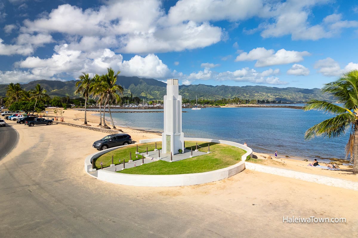pulled back view of haleiwa beach park and war memorial, plus a family on the beach and cars in the parking lot