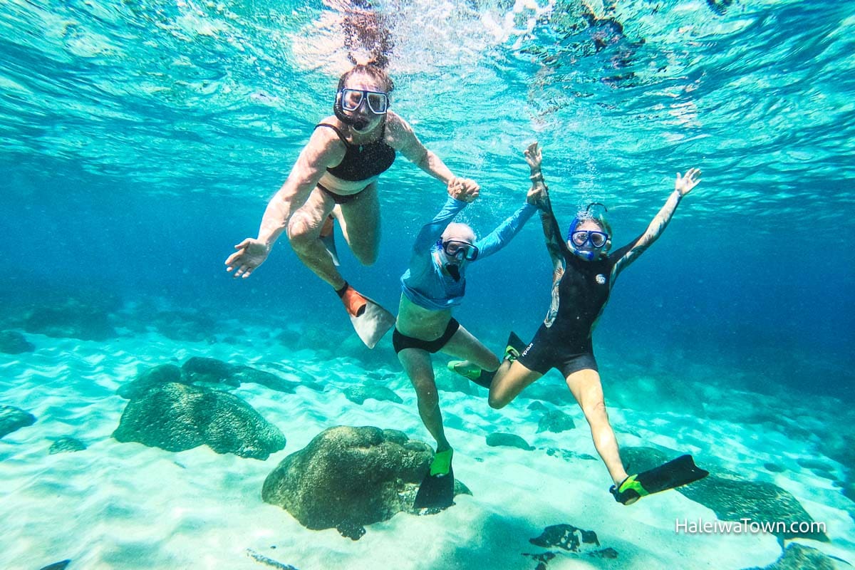 two women and one older man holding hands underwater snorkeling in clear blue ocean