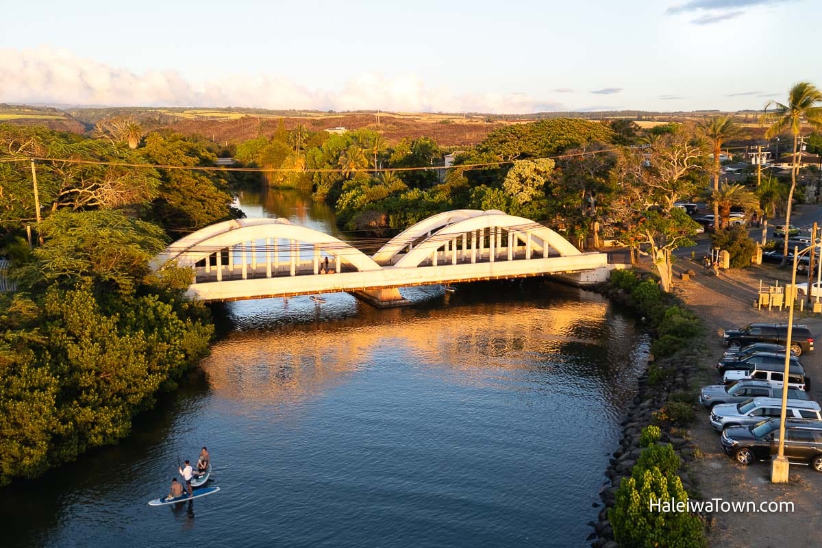 aerial view of Haleiwa Rainbow Bridge lit up during sunset and some standup paddleboarders on the river