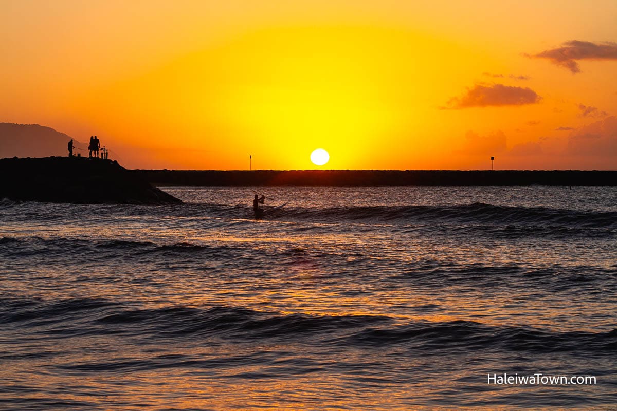 sun setting behind the rocks at the haleiwa boat harbor, a kayak man paddling across the channel and a few people on the rocks watching the sunset