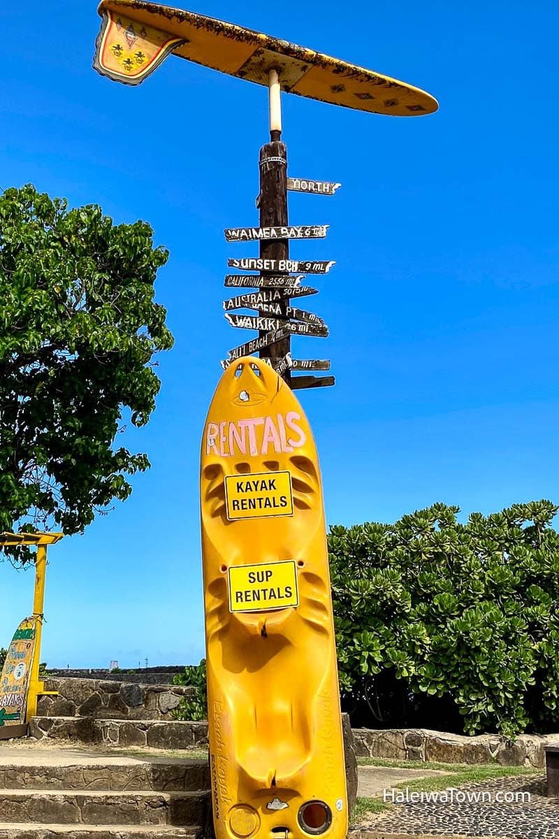 wooden post with a large surfboard on top and another board as the main sign for a rental shop offering kayaks and sup or stand-up paddleboards