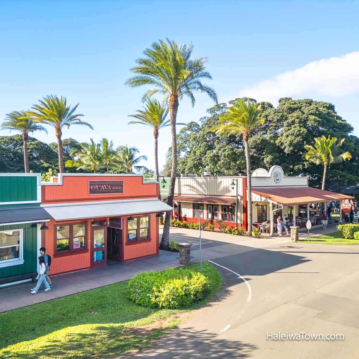 center of haleiwa town with a green, red, and tan building with restaurants and shops, palm trees and people on the sidewalks