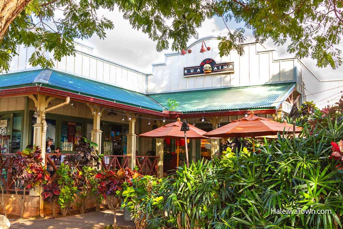 tan vintage building with blue metal roof and a sign for kuaaina burgers surrounded by plants in front and outdoor seating with red umbrella for shade