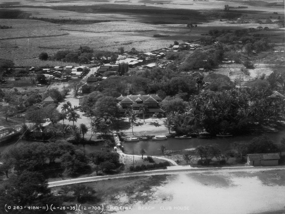 aerial historic view of haleiwa beach club house, railroad tracks, wooden bridge, and boats in the anahulu stream in 1935 surrounded by palm trees