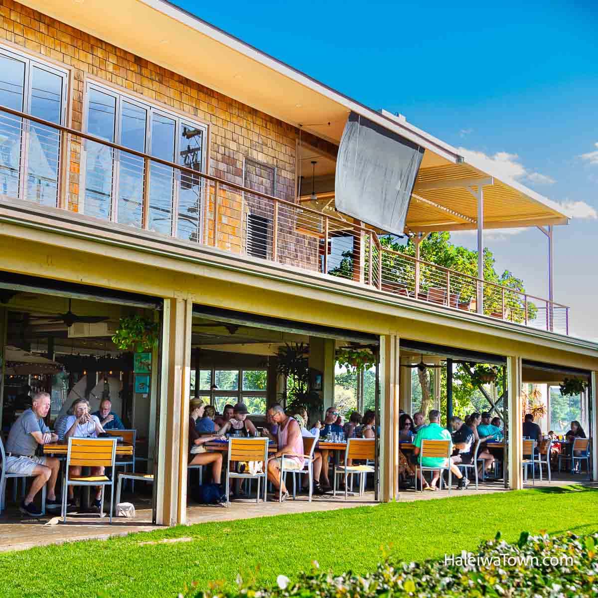 two story building restaurant with covered outdoor seating full of people dining overlooking green grass on a sunny day