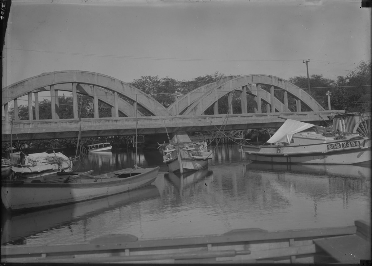 bridge with large arches over the anahulu stream with several fishing boats attached to it by thick ropes