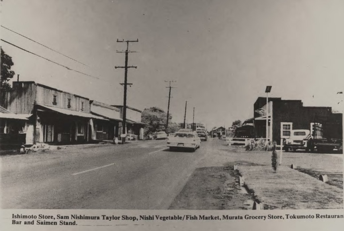 historic black and white view of haleiwa town's two way street with cars driving by and stores on both side of the street