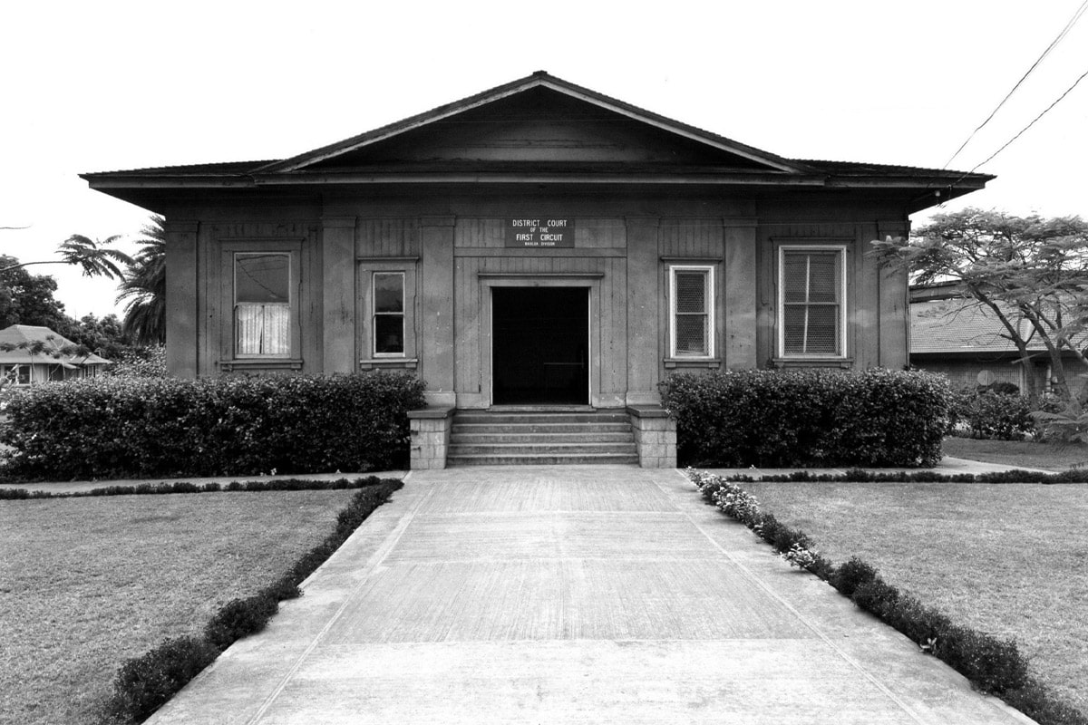 historic view of Waialua Court House building with double doors opened windows on both sides and a pitched roof in the middle with a long concrete driveway in front