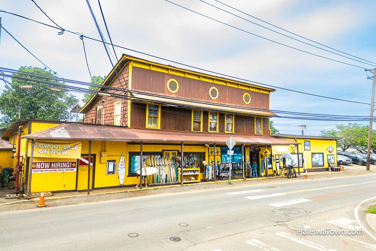historic brown and yellow wooden vintage building with surfboards and clothing outside for sale