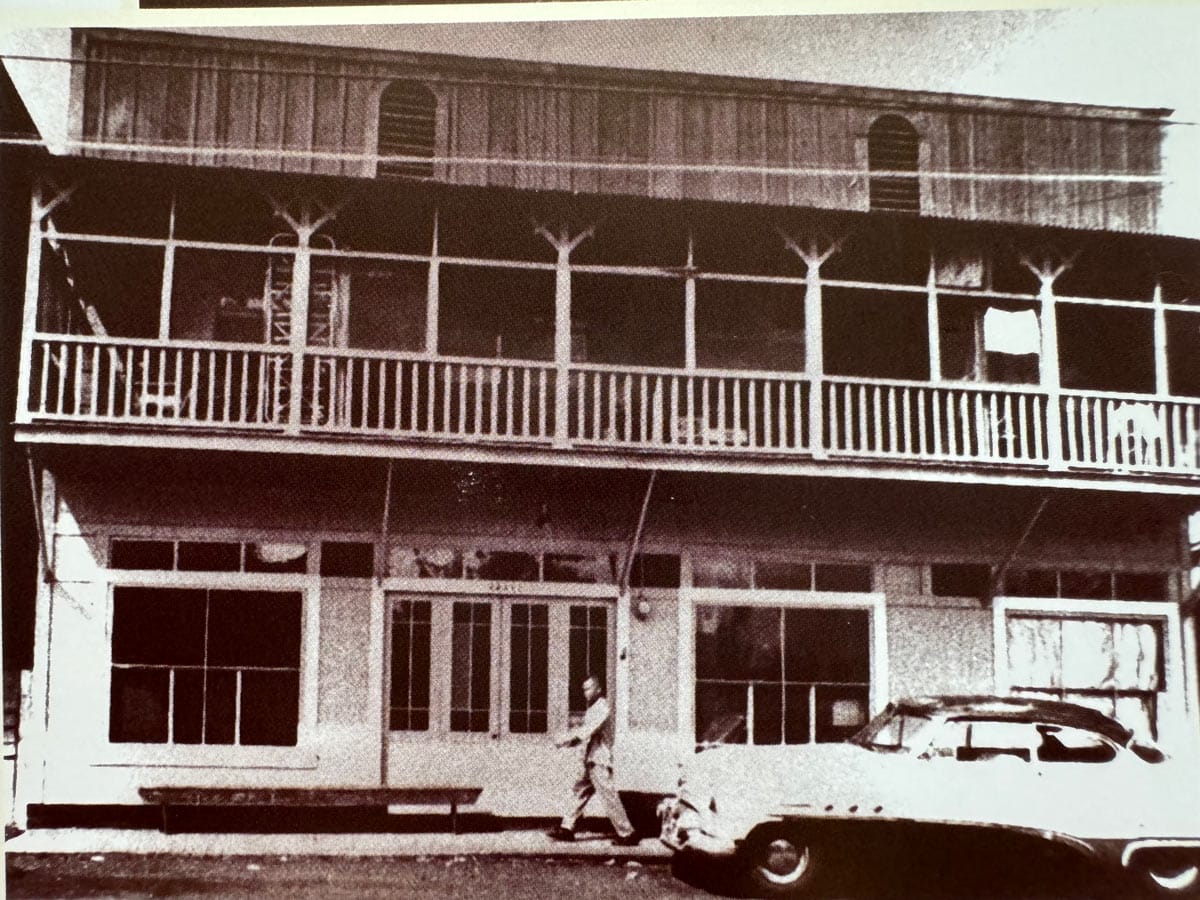 historic blac and white view of wooden two story building with a deck on the second floor and an old car parked in front
