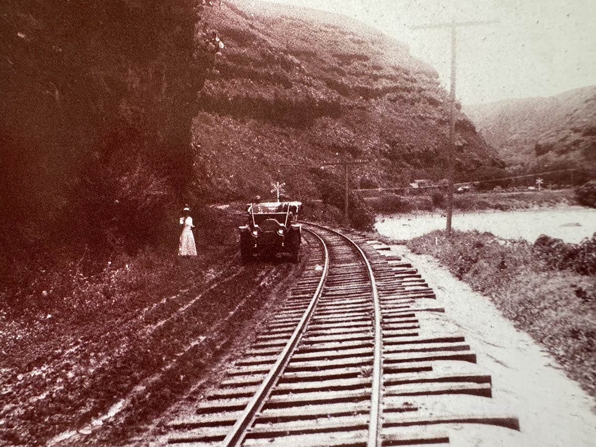 old railroad line surrounded by tall mountains and the ocean with a car and a women standing next to it