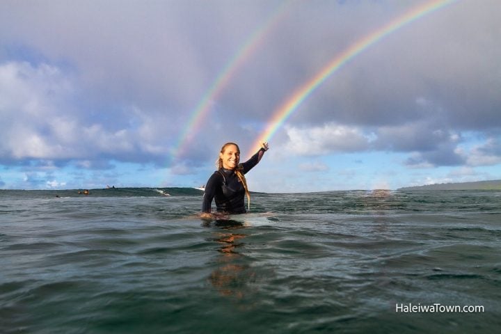 girl sitting on surfboard in the ocean pointing to rainbow in the sky