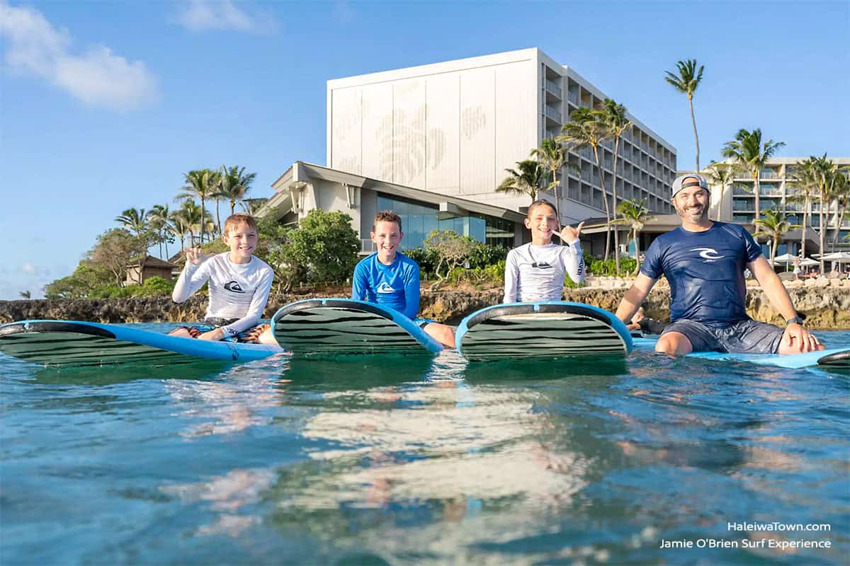 kids and surf instructor in the ocean sitting on a surfboard in front of turtle bay resort